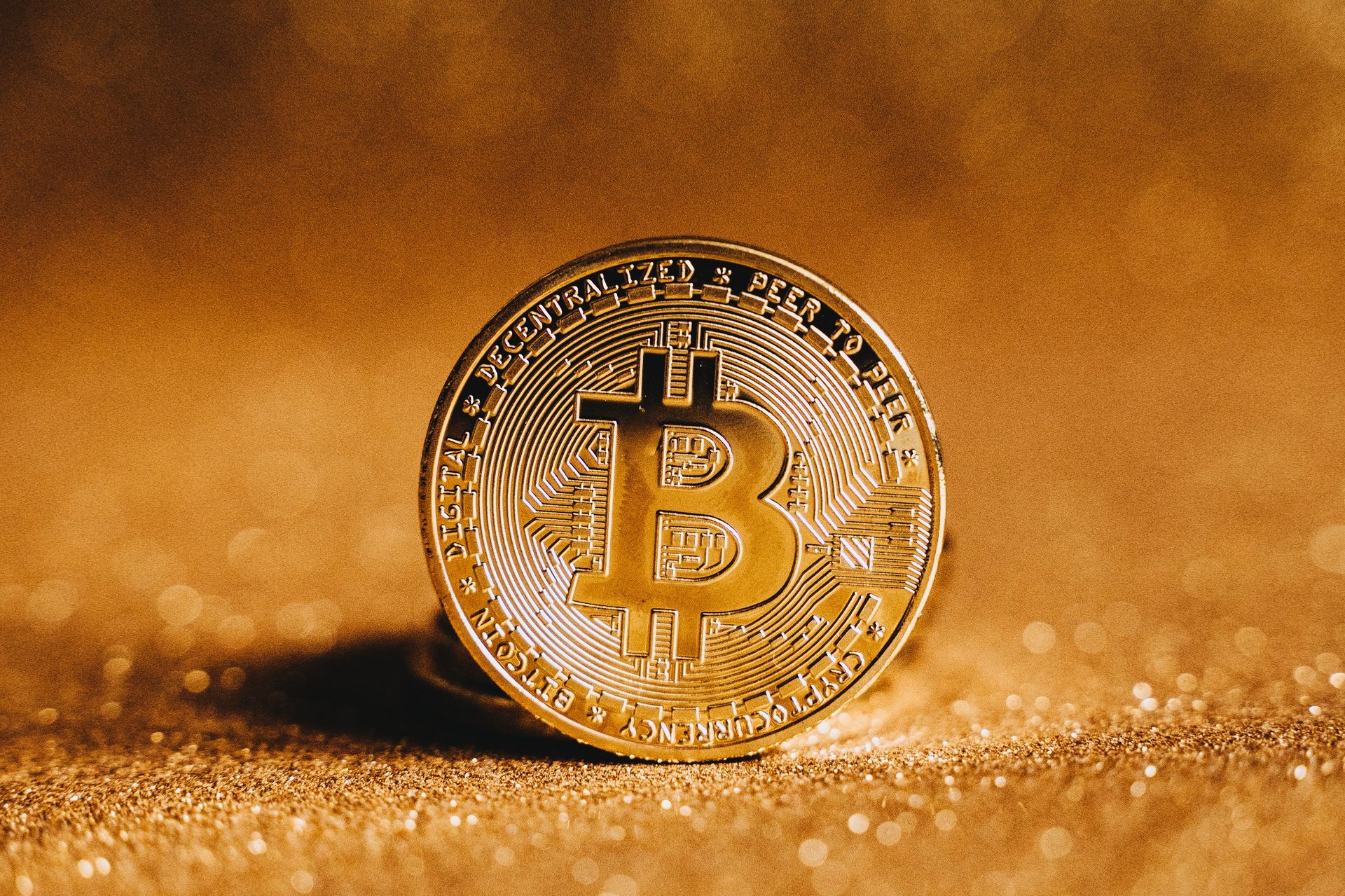 Nahaufnahme einer physischen Bitcoin-Münze mit sichtbarem B-Symbol auf goldfarbenem, glitzerndem Untergrund