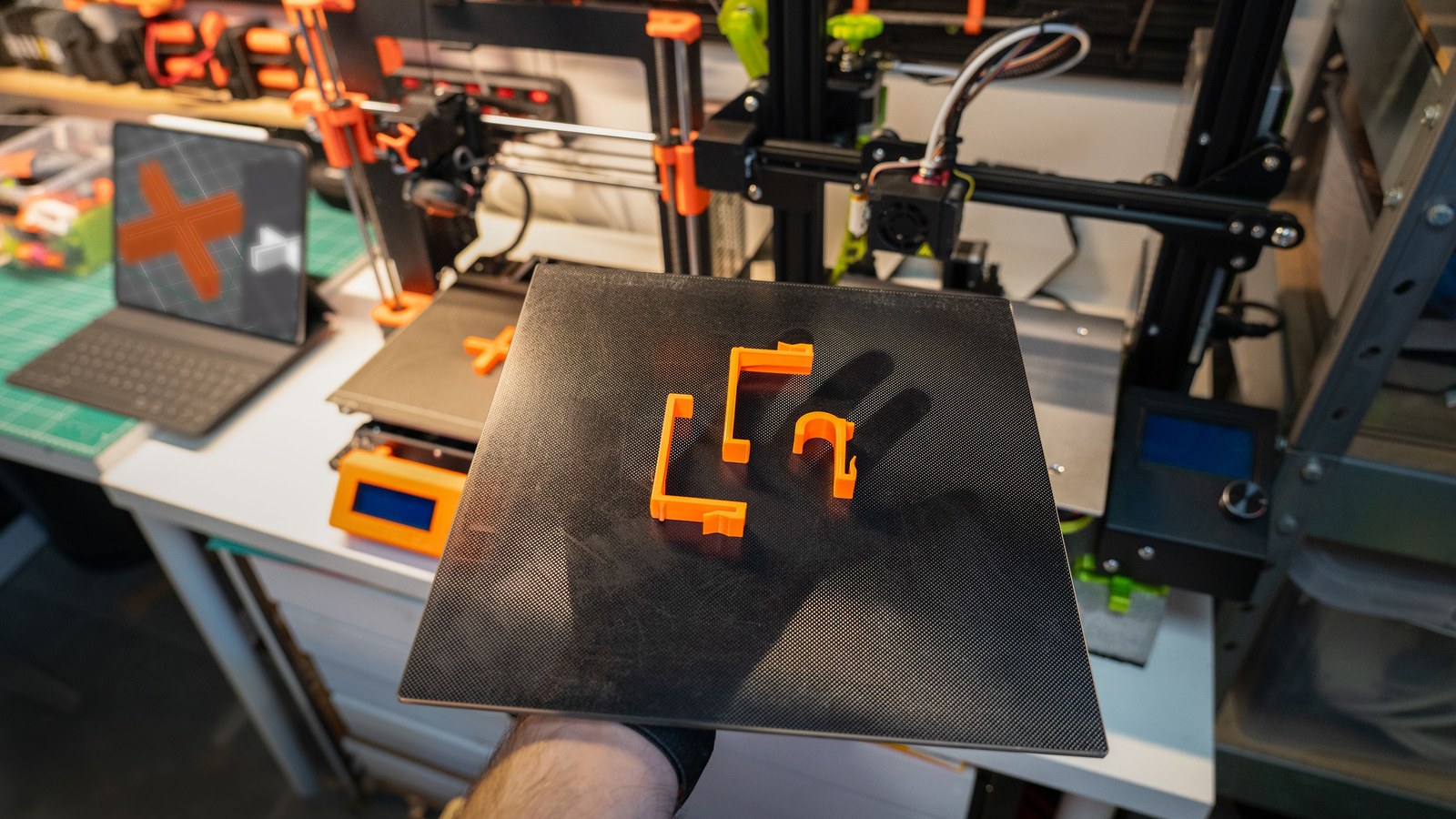 Hand hält eine Druckplatte mit mehreren selbst gedruckten orangefarbenen Halterungen vor einem FDM-3D-Drucker