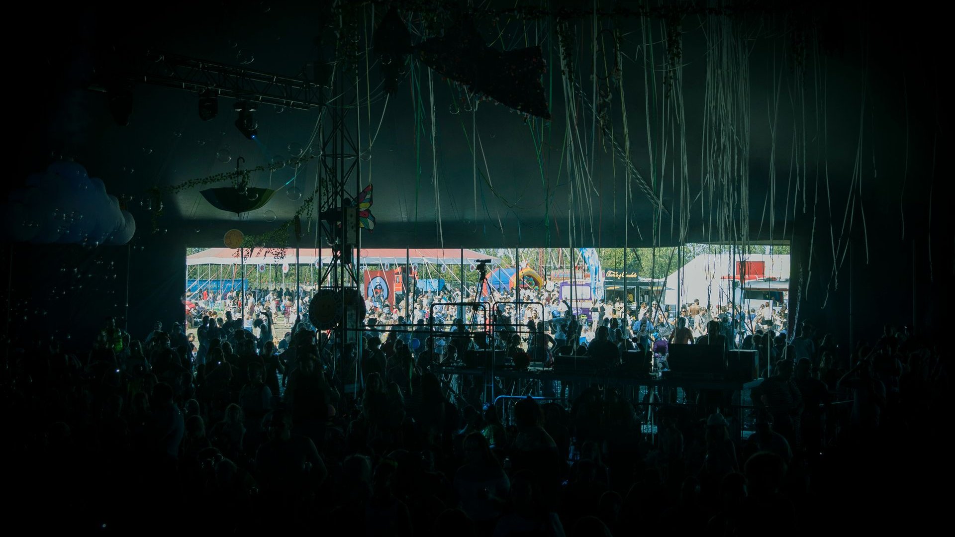 Silhouetten von Festivalbesuchern in einem dunklen Zelt, im Hintergrund ein sonniges Festivalgelände (Symbolbild)